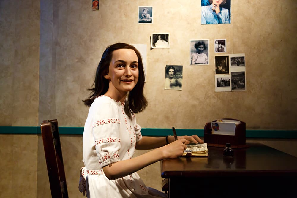 Una figura de cera de una mujer sentada ante un escritorio con lápiz y papel, rodeada de fotografías en la pared.