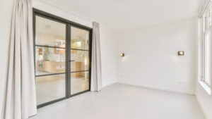 A bright, empty room with white walls, floor-to-ceiling curtains, and sliding glass doors leading to an adjacent room.