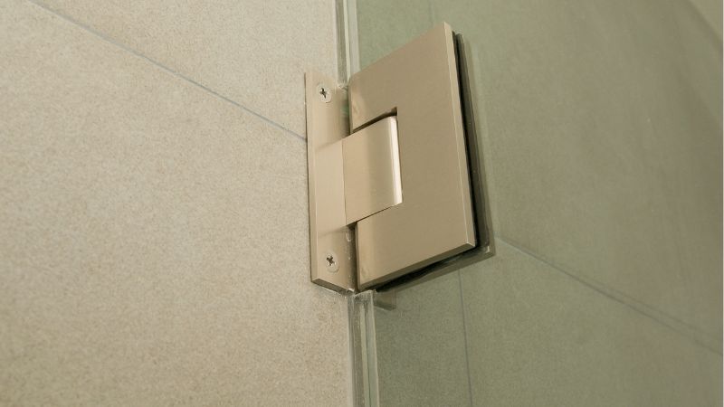 A close-up of a stainless steel bathroom stall hinge attached to a frosted glass panel.