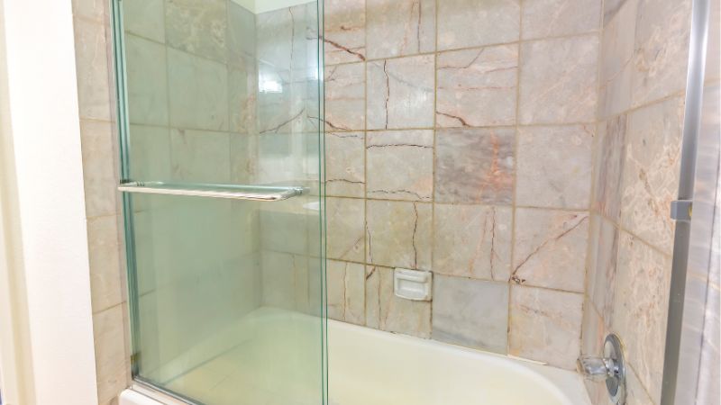 Cuarto de baño moderno con cabina de ducha de cristal y bañera blanca, con azulejos de mármol marrón claro en las paredes.