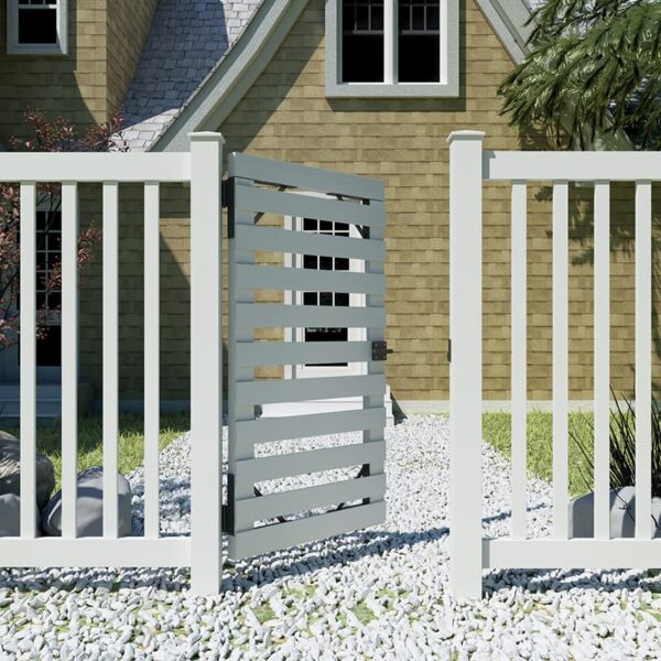 A white picket fence with a slatted gate and Double Side Gate Latch stands before a tan house with a gray roof. Gravel lies near the fence.