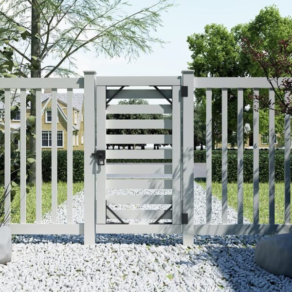 A white slatted fence with a lockable gate stands on a pebbled path, bordered by greenery under a clear blue sky.