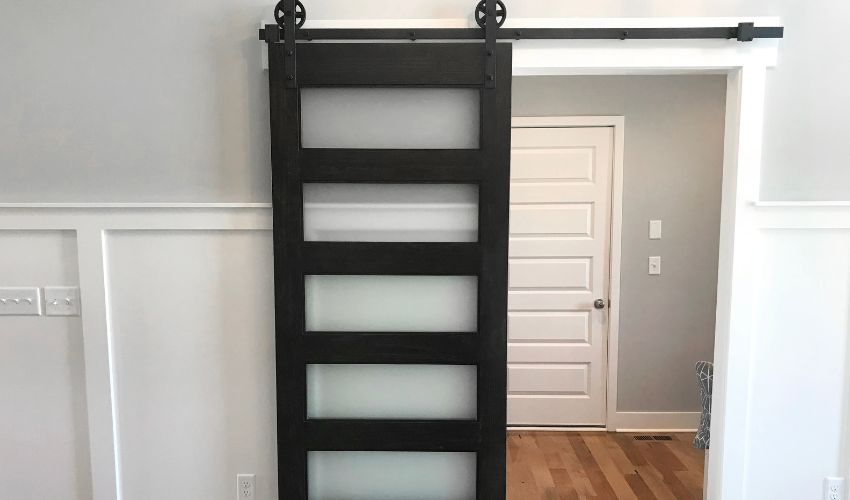 A black barn door with frosted glass reveals a white four-panel door in a gray-and-white room with wood flooring, blending style and function.