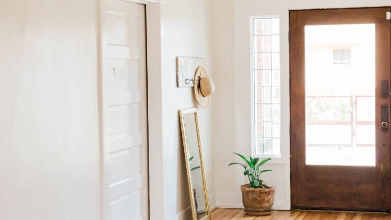 Un couloir lumineux présente une porte en bois fabriquée, avec un chapeau de paille sur un crochet, un miroir penché et une plante en pot présentant les tendances de design 2024.