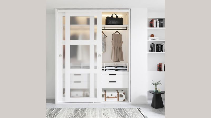 A white wardrobe with sliding doors houses clothes, bags, and luggage. Nearby, shelves hold books and decor; a black table sits by a plant.