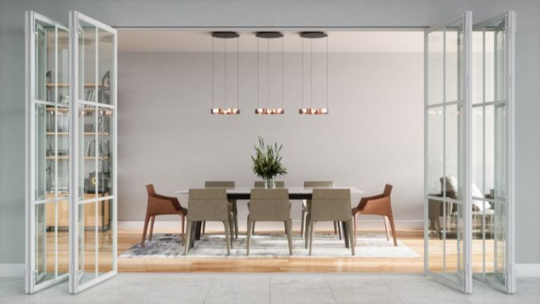 Salle à manger moderne avec une grande table, huit chaises, une plante centrale. Luminaires suspendus au-dessus. Portes vitrées ouvertes et étagères décoratives visibles.