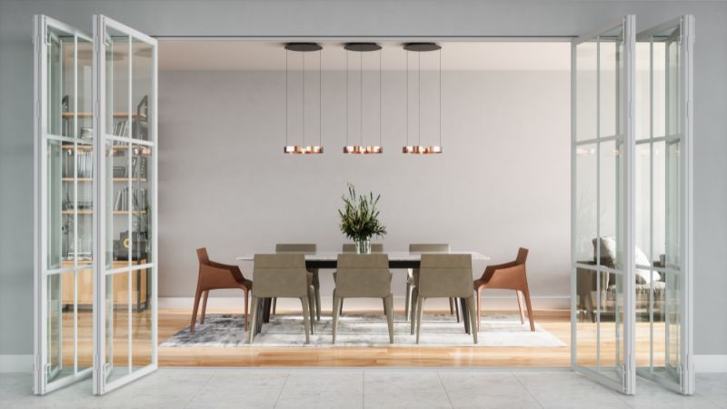 Modern dining room with a large table, eight chairs, plant centerpiece. Pendant lights above. Open glass doors and decor shelves visible.