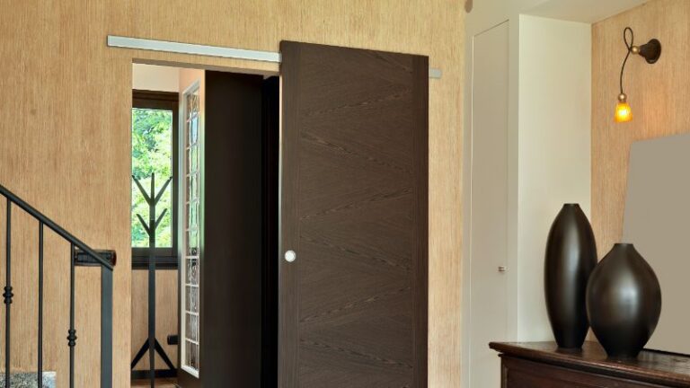 Une pièce avec une porte coulissante en bois partiellement ouverte présente les tendances de design 2025. À proximité, découvrez un porte-manteau, des balustrades, une commode avec des vases et une applique murale élégante.