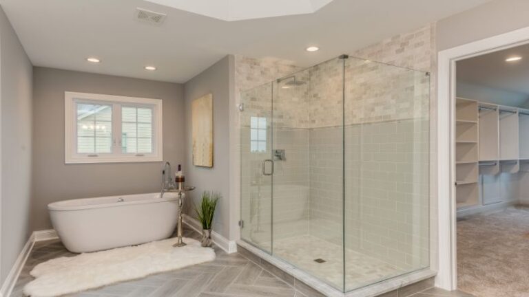 Une salle de bain moderne dispose d'une baignoire îlot, d'une douche élégante avec porte vitrée, d'un élégant carrelage gris et d'une fenêtre. Un tapis et un dressing sont visibles.