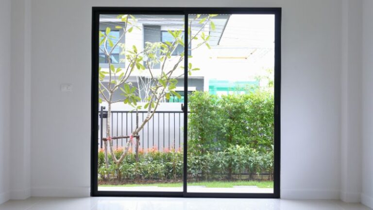 Une élégante porte coulissante en verre à cadre noir complète le jardin avec des arbustes, un arbre et une élégante clôture métallique en arrière-plan.