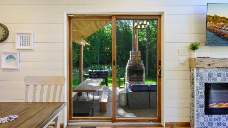 Vue à travers les 10 premières portes coulissantes montrant un patio couvert avec table de pique-nique, barbecue, cheminée parmi les arbres ; coin repas à l'intérieur.