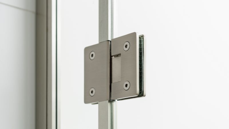 Närbild av metallgångjärn som förbinder glaspaneler, troligen från en modern duschdörr, mot en vit bakgrund.