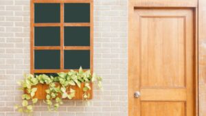 Una ventana con marco de madera, con una jardinera y enredaderas, se encuentra junto a elegantes puertas de madera en una pared de ladrillos claros.