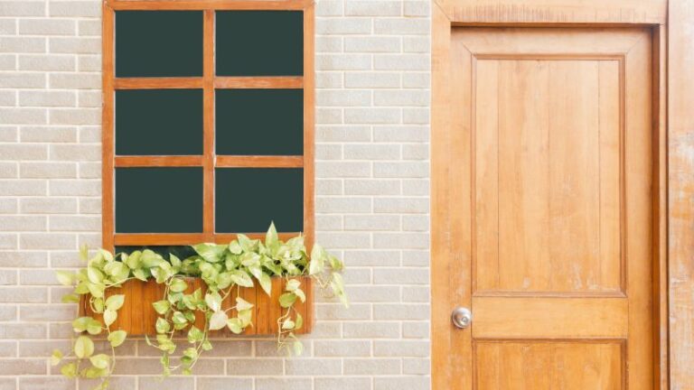 Une fenêtre à cadre en bois avec une jardinière et des vignes se trouve à côté d'élégantes portes en bois sur un mur de briques claires.