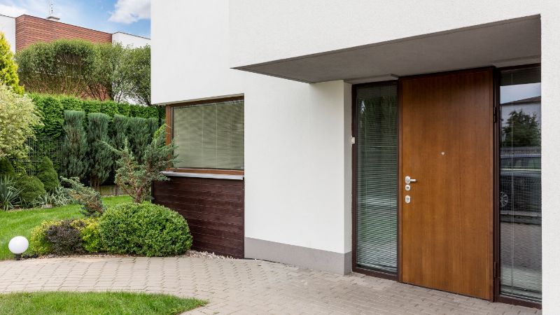 Entrada de casa moderna con puerta de madera, panel lateral de vidrio, paredes blancas y vegetación ajardinada junto a un camino pavimentado.