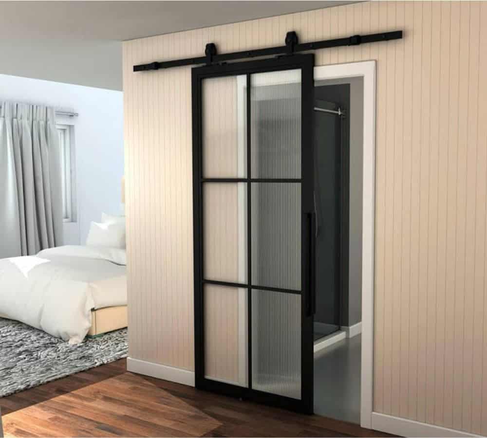Una puerta de granero corrediza con marco de metal negro y vidrio esmerilado separa un dormitorio y un baño, mostrando su estilo elegante y moderno.