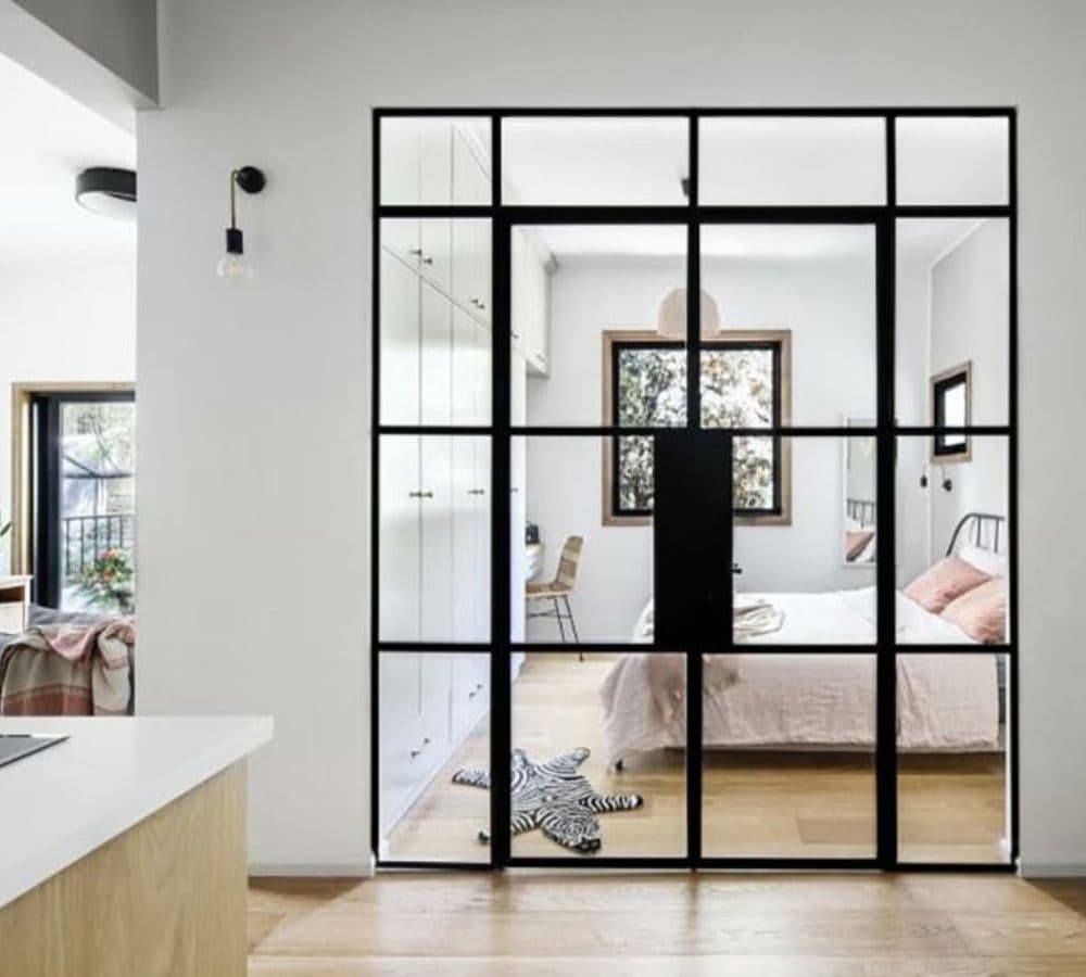 Dormitorio con paredes blancas, puertas de vidrio con marco de metal, cama, escritorio, ventana y una alfombra de cebra sobre piso de madera.