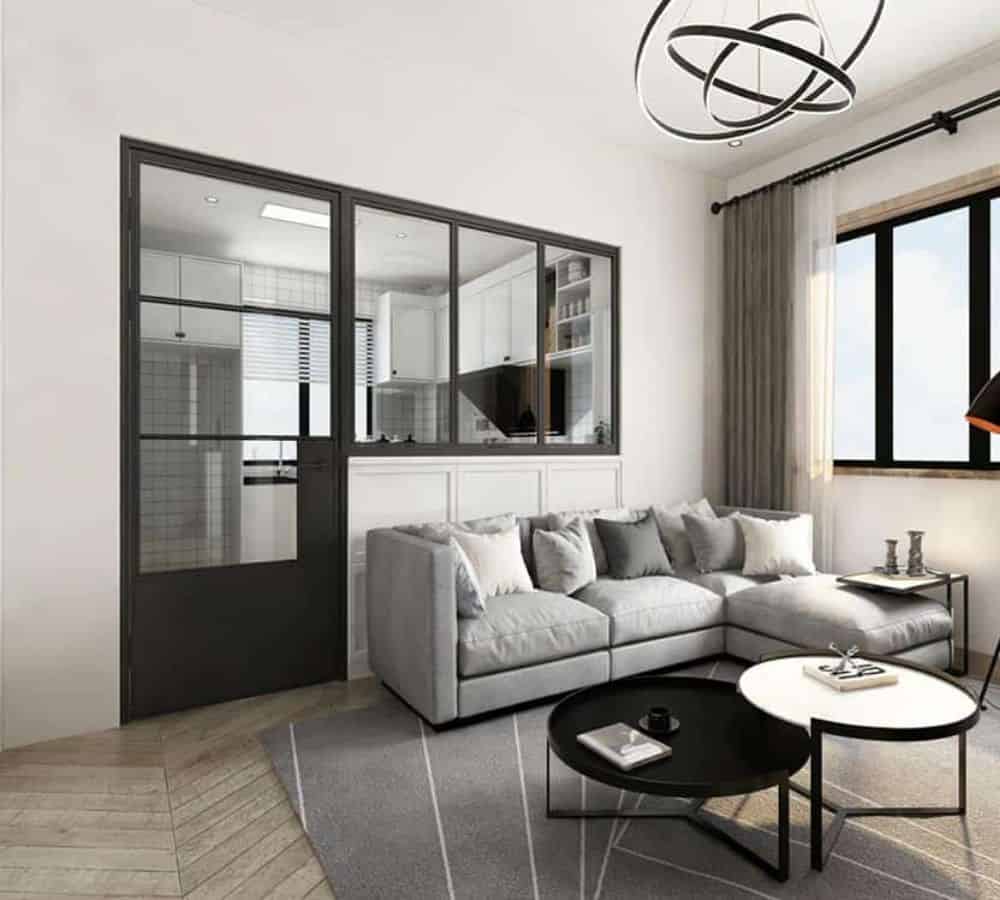 Moderna sala de estar con sofá seccional gris, mesas redondas, luz de techo y puertas de vidrio con marco de metal que dividen la cocina.
