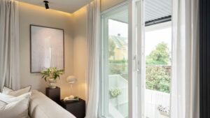 Luxurious bedroom featuring large glass sliding doors opening to a cozy balcony with lush greenery, elegant white curtains, and contemporary decor, perfect for a relaxing home environment.