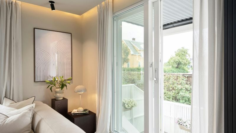 Luxurious bedroom featuring large glass sliding doors opening to a cozy balcony with lush greenery, elegant white curtains, and contemporary decor, perfect for a relaxing home environment.