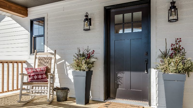 Stylish black front door with window panes, flanked by outdoor lanterns and potted plants, on a white exterior wall, perfect for American home entrances.